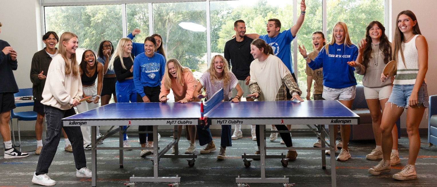 A group of college students cheering and laughing around a blue ping-pong table during a lively indoor game.